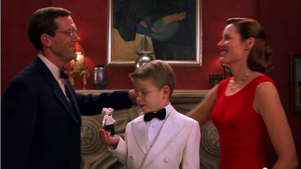 The image shows a scene from the movie Stuart Little, with a man in a black suit and a woman in a red dress on either side of a boy in a white tuxedo with a black bowtie holding a clothed mouse in his hand. In the background can be seen Berény's 